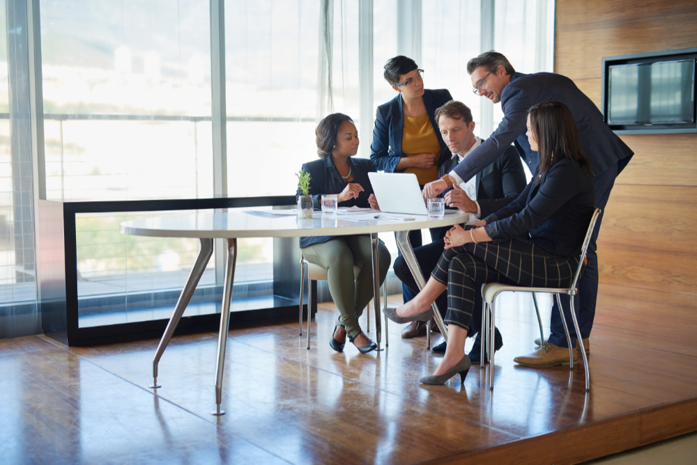 Five business people stand over a laptop on a table in a conference room to discuss marketing strategy best practices for asset management firms